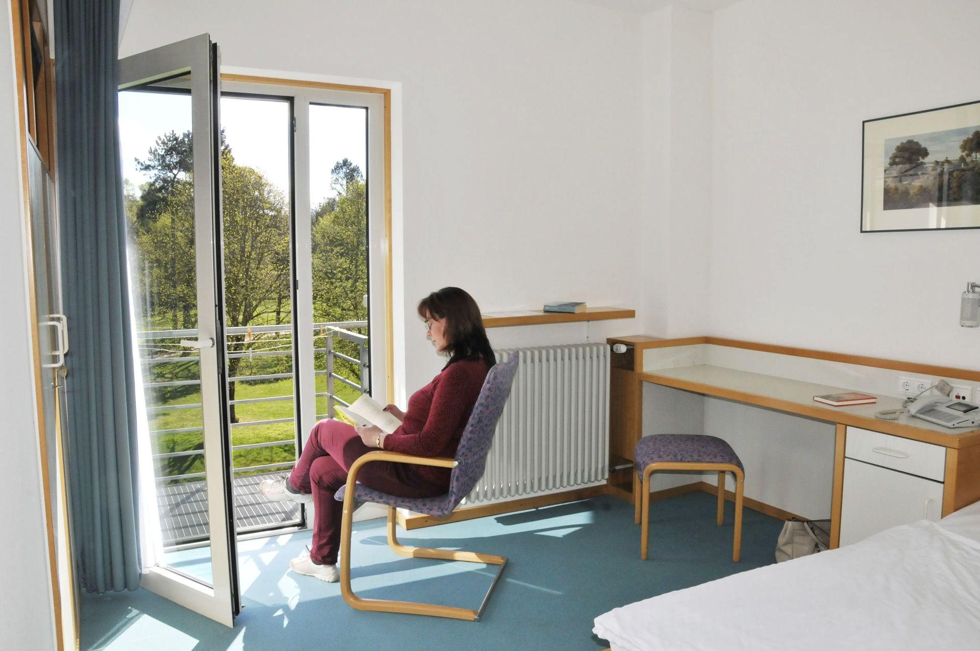 Eine Patientin sitzt in ihrem Zimmer mit Blick nach draußen durch die geöffnete Balkontüre. 